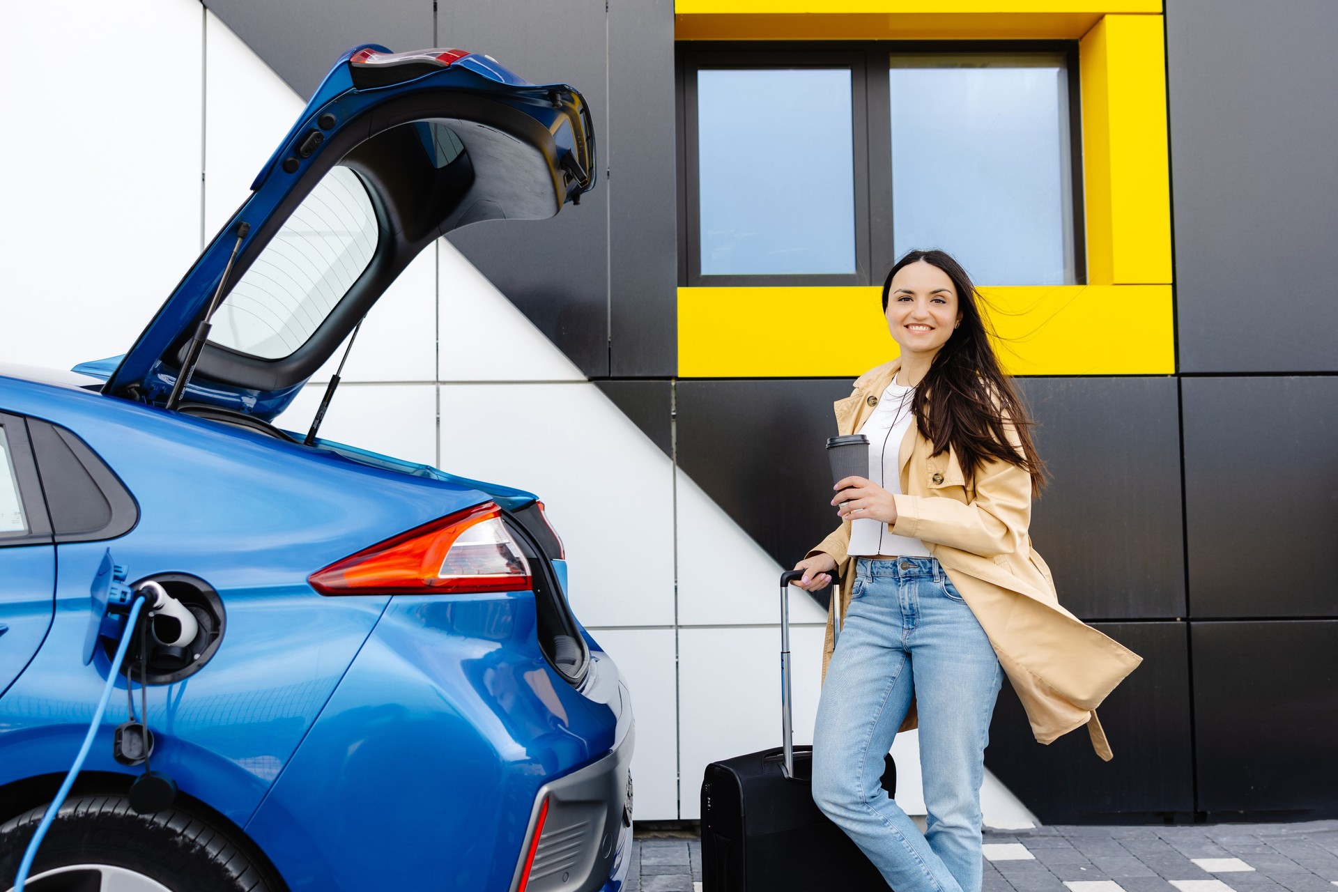 Young woman with cup of coffee waiting while her electric car charging, sustainable and economic transportation concept
